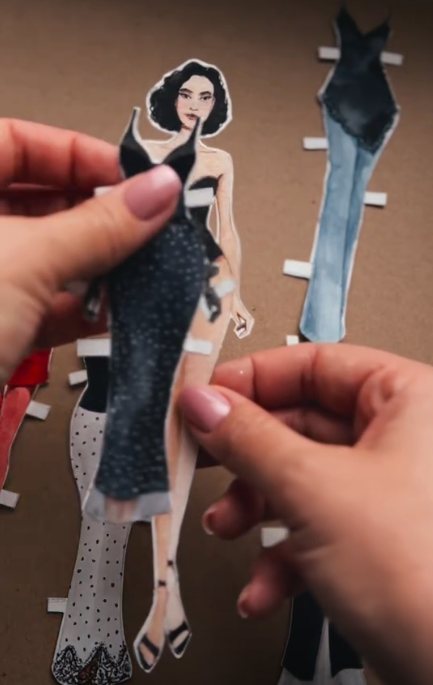 Mão encaixando um vestido preto de papel na boneca
