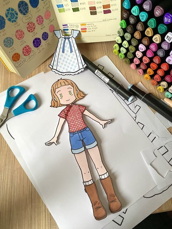 Boneca de papel com vestido azul, tesoura e canetinhas coloridas sobre a mesa