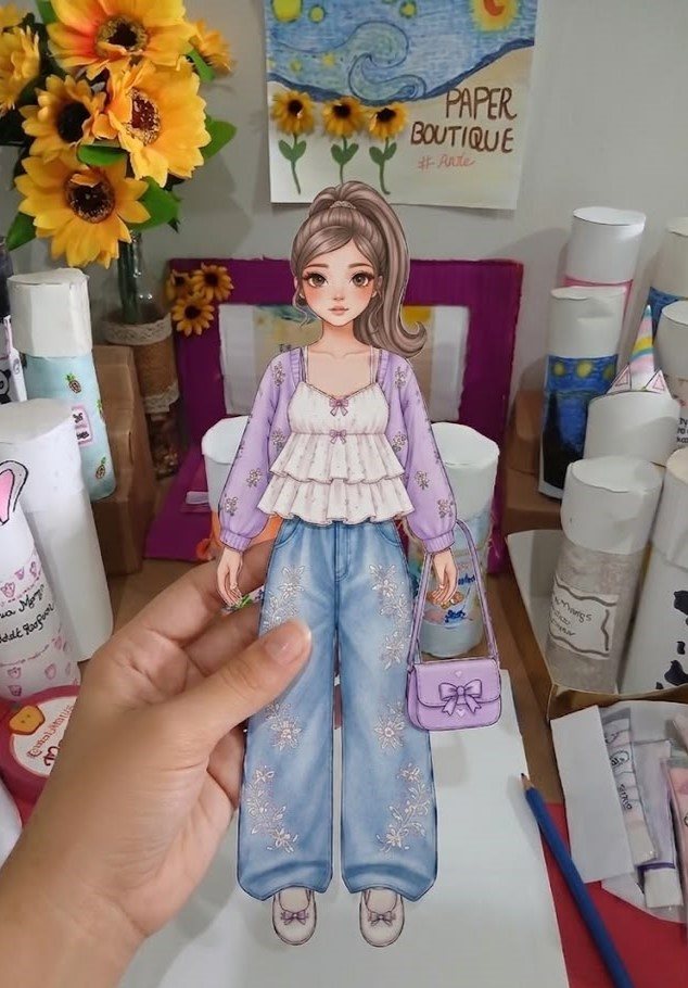 Mão segurando boneca de papel moderna montada com casaco lilás, calça jeans e bolsa